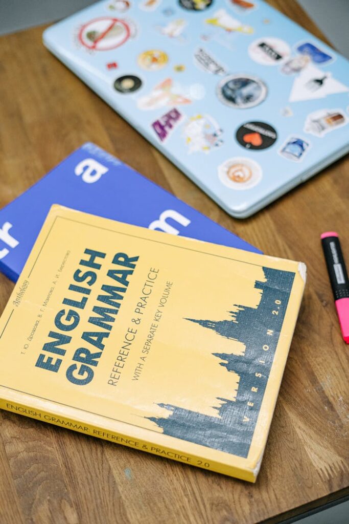 pexels photo 5676740 High-angle view of an English grammar book and a laptop with stickers on a wooden desk.